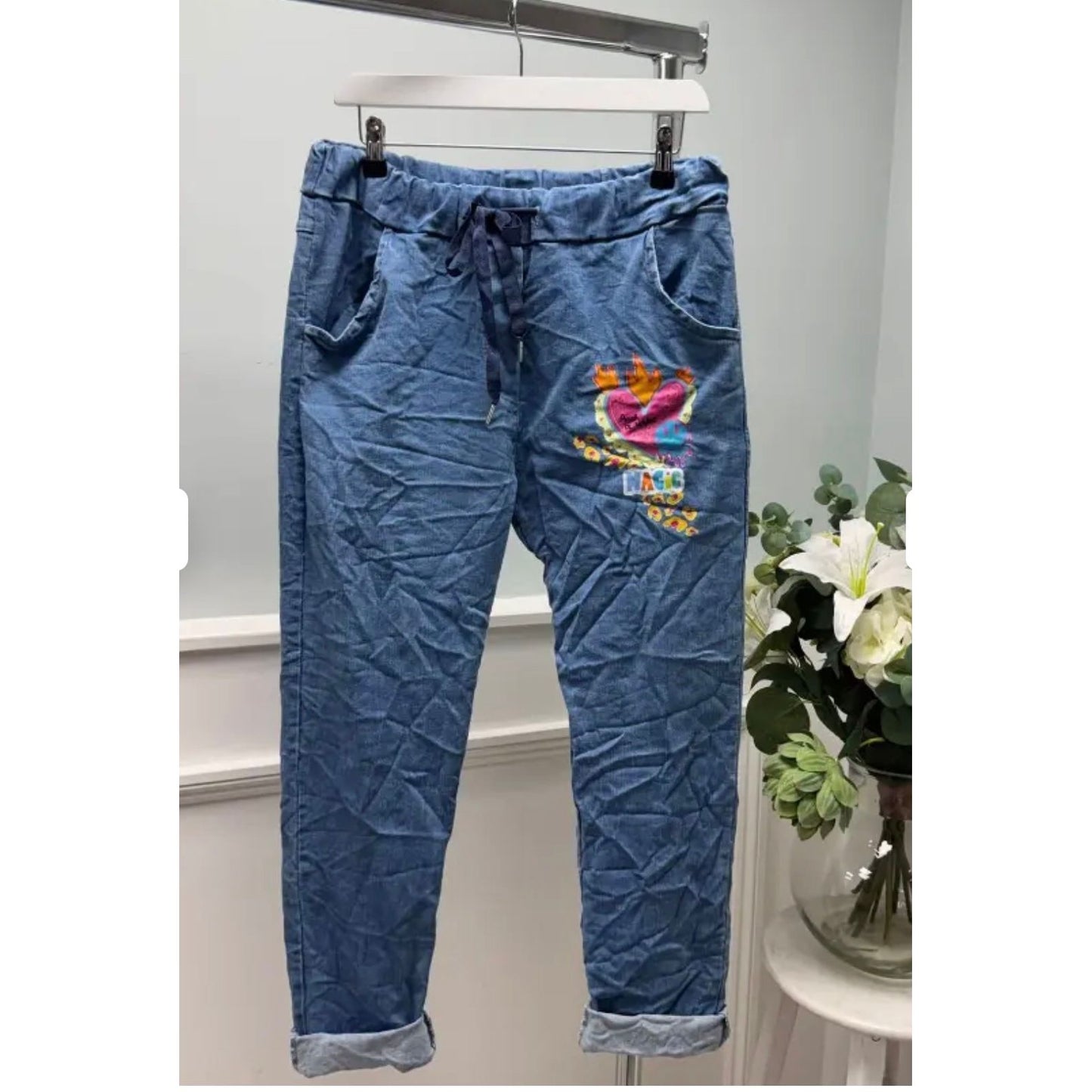 Blue denim jogger pants with colorful graphic on a hanger against a white background.