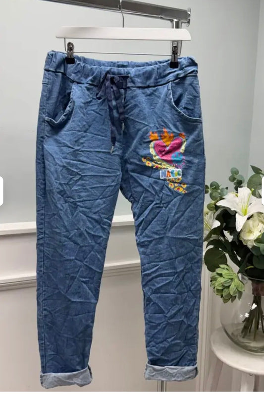 Blue denim jogger pants with colorful graphic on a hanger against a white background.